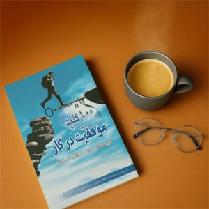 تصویری از کتاب «۱۰۰ کلید موفقیت در کار» که روی یک میز قهوه‌ای در کنار یک فنجان قهوه داغ و یک عینک قرار گرفته است.